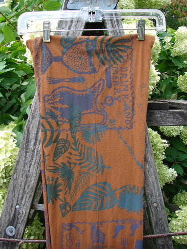 Barclay Curly Edge Center Seam Scarf Wrap in Fall Forest Friend rust, an OSFA scarf with a center seam and softly curled edges. The lightweight fabric shows a deep rust base printed with blue-green botanical and fruit motifs, hanging from clips on a wooden ladder amid greenery.