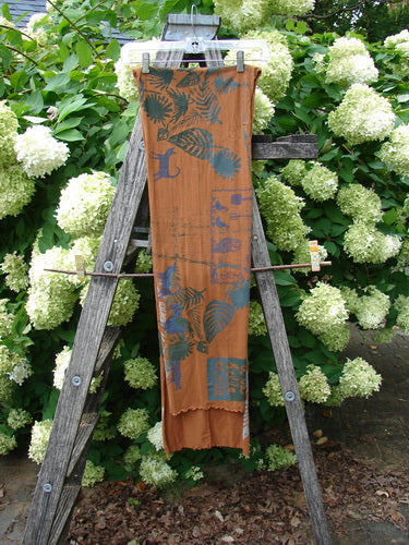 Barclay Curly Edge Center Seam Scarf Wrap in Fall Forest Friend Rust, an elongated rust-orange wrap with teal fern and leaf motifs and a curly edge. It hangs vertically from a wooden ladder-style display, with a center seam and soft drape.