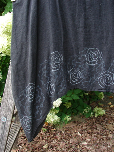 Barclay Linen Pinafore Curve Drop Side in black, Size 2, with a soft linen weave and pale rose border motifs tracing the lower hem. The fabric hangs in a loose, asymmetrical drape outdoors, its dark surface set against green foliage and mulch.