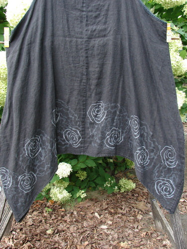 Barclay Linen Pinafore Curve Drop Side Rose Border in black, size 2, with a draped linen silhouette and curved drop sides. Pale rose border motifs trace the lower hem, while the dark fabric hangs in soft folds against a leafy outdoor backdrop.