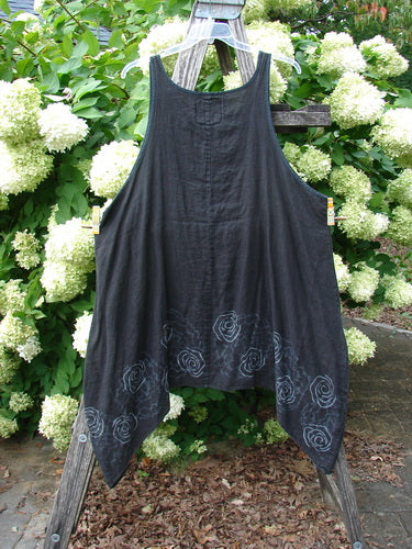Barclay Linen Pinafore Curve Drop Side Rose Border in black, size 2, shown as a sleeveless linen pinafore with a loose, A-line drape and curved drop sides. Embroidered rose borders trace the hem, while the dark fabric hangs from a wooden frame against dense white hydrangeas and green foliage.