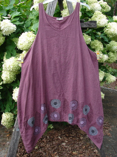 Barclay Linen flared pinafore shift dress in berry, size 2, with a sleeveless scoop neckline and wide draped silhouette. The linen fabric falls in soft folds, and circular pinwheel turn leaf motifs line the pointed hem, photographed hanging outdoors against leafy greenery and white blooms.