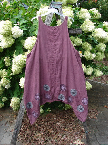 Barclay Linen Flared Pinafore Shift Dress in berry, size 2, hangs from a rustic wooden ladder with a soft, sleeveless A-line shape and wide shoulder straps. The linen fabric has a matte, gently wrinkled texture, and the hem is decorated with circular pinwheel turn leaf motifs in muted gray against the mauve-purple body.