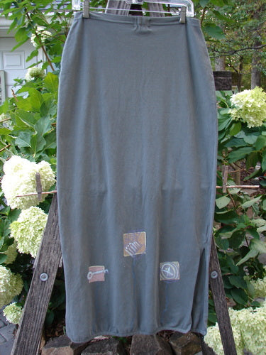 2001 Slim Skirt Junk Drawer Kelp Size 0 displayed draped over a wooden ladder, showcasing its long, elegant design and flattering full flattened waistline with side vents and a pegged hemline.