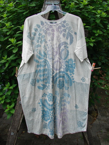 Barclay Long Tee Tie Back Dress Botanical Garden Rain Cream OSFA features a seamless front and back bodice, rounded neckline, dual pockets, and dolman sleeves, displayed on a clothes rack.