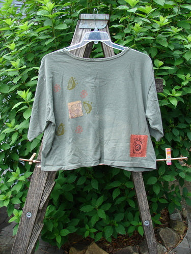 1991 Short Sleeved Crop Tee Fish Flower Olive Drab OSFA displayed on a hanger, featuring vintage double fish and continuous flower design, rolled neckline, and slight rear center fade, highlighting BlueFishFinder's vintage style.