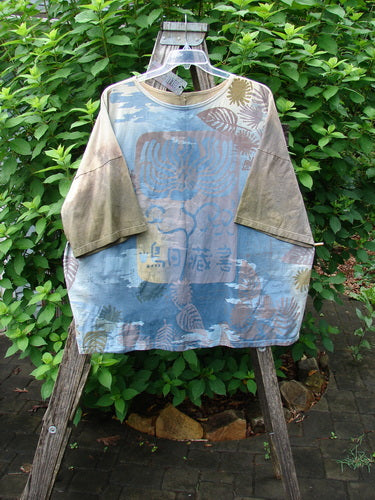 Barclay NWT Center Seam Wide Line Tee Earth & Sea Mottled Lens OSFA, featuring a rounded neckline, drop shoulders, and vertical center seam, displayed in an outdoor setting amidst plants and artistic elements.