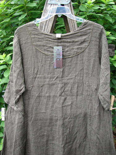 Barclay NWT Linen Cross Over Urchin Pocket Dress Unpainted Earth Size 1 displayed on a wooden swinger, showcasing its crossover V-neckline, drop shoulders, and double side pockets in a lightweight linen fabric.