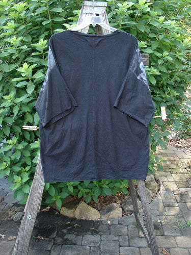 Barclay Raw Edge Flutter Collar High Low Top Dogwood Shadow Black OSFA hanging on a wooden rack, showcasing its rounded flutter neckline, raw edge dolman sleeves, and distinctive high-low hemline.