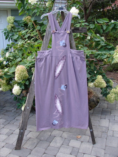 1996 Souvenir Jumper Turn Path Petunia Size OSFA displayed on a wooden ladder, featuring a full gathered skirt, criss-cross back, thick straps, and an abstract painted bib. Made from organic cotton.