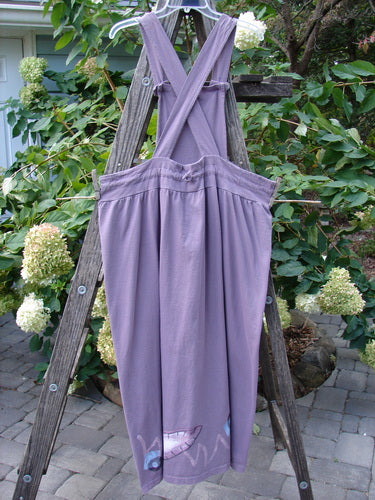 1996 Souvenir Jumper Turn Path Petunia Size OSFA displayed on a wooden ladder, featuring a gathered skirt, crisscross back, straight straps, and abstract painted bib, with organic cotton fabric.