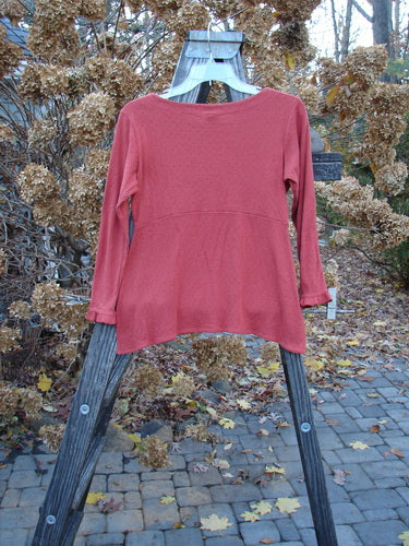 2000 Wool Pointelle Anais Top Unpainted Bittersweet Tiny Size 1: A red shirt on a wooden stand, with sweet lettuce edging, empire waist seam, and tailored waistline. Cozy and breathable, made from soft merino wool.