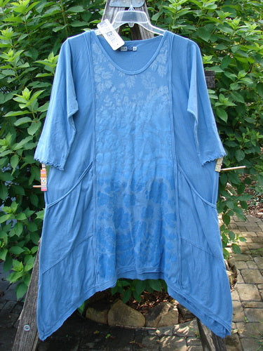 Barclay NWT Wildflower Tunic Garden Blend Blue Bird Size 0 displayed on a hanger, highlighting its raw-edged front, flutter hemline, and exterior pockets with three-quarter sleeves amidst a garden-themed backdrop.