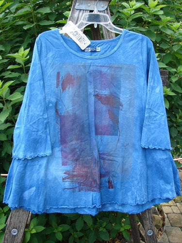 Barclay NWT Three Quarter Sleeved Cafe Tee in Shadow Blend Mottled Blue Sky, displayed outdoors on a hanger, showcasing its unique A-line shape, rounded neckline, and curly edgings.