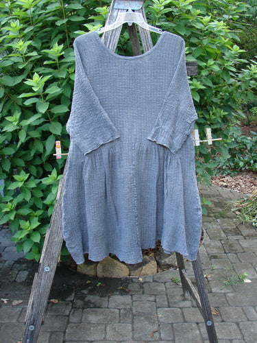 Barclay Crinkle Gauze Two Pocket Dolly Dress Mottled Slate Size 1 on a wooden rack, showcasing its flouncy dual-pocketed skirt, downward curved empire waist, and three-quarter sleeves with a varying hemline.