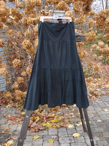 Barclay Batiste Fold Over Wave Layer Skirt Unpainted Black Size 2 displayed on a wooden clothes rack, highlighting its full paneled waistline and varying hemline with horizontally pinched waves.