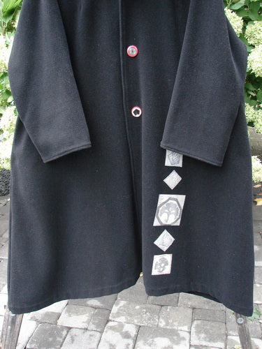 1999 Patched Woven Wool Overcoat in jet black, size 2, with a long straight silhouette, wide sleeves, and a soft woven wool texture. A row of small front buttons and a vertical patch panel with pale geometric appliqués add contrast against the dark coat.