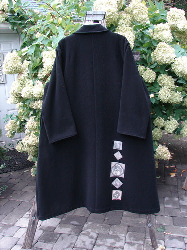 1999 Patched Woven Wool Overcoat in jet black, size 2, with a long, loose silhouette, wide sleeves, and a high collar. The woven wool body falls in a smooth, heavy drape, while a vertical row of small patched squares accents the lower front.