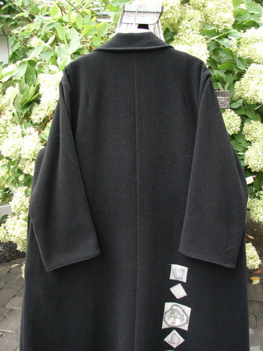 1999 Patched Woven Wool Overcoat in jet black, size 2, with a long straight silhouette, wide sleeves, and a soft woven wool texture. Small patch details trail near the lower back, and the coat hangs outdoors against green foliage.