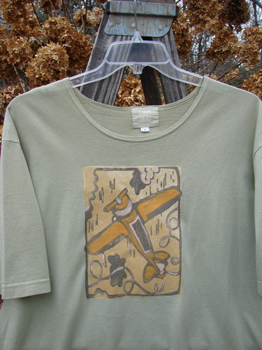 2001 Short Sleeved Tee Glider Plane Kelp Size 1 displayed on a hanger, featuring a flattened neckline, drop shoulders, wide short sleeves, and a glider plane theme painted on mid-weight organic cotton.