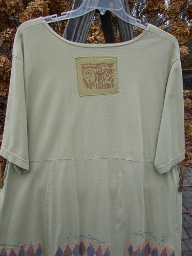 Image: A shirt with a logo on it, featuring a square patch with text. The shirt is on a hanger, with a close-up of a plastic object.

Alt Text: 1993 3 Square Dress Trinket Diamond Kelp OSFA: A vintage Blue Fish shirt with logo and patch, showcasing a unique design with sectional panels and a slight empire waist seam.