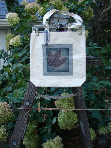 1998 PMU Patched Organic Cotton Canvas Tote Bag featuring purple oak leaf patches, displayed on a wooden ladder. The tote has long shoulder straps, a wide top opening, and a gusseted bottom.