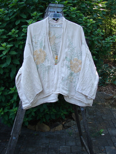 Magnolia Pearl Linen Embroidered Claudio Kimono Lilac Water OSFA displayed outdoors, showcasing its wide, swingy three-quarter sleeves, dolman shoulders, and pastel floral embroidery on a medium weight linen fabric.