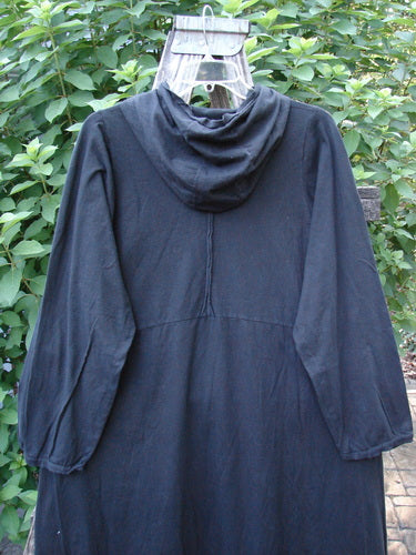 Barclay NWT Hi Low Hooded Pocket Tunic Unpainted Black Size 0 displayed hanging on a wooden pole, showcasing its cordless hood, raw-edged sleeves, and flared swing design with dual front pockets.