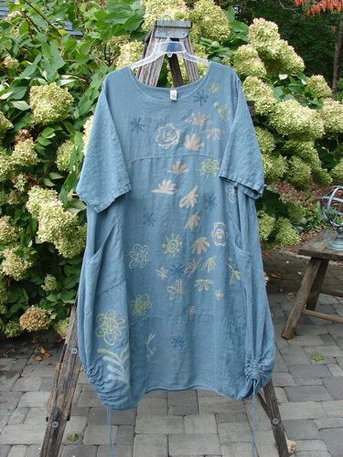 Barclay Linen Lucky Urchin Dress Floral Drift Delaware size 2 in faded blue linen, hanging on a wooden rack. The loose, short-sleeve silhouette shows a soft drape, floral and shell-like prints in pale yellow and blue, and gathered hem details against lush garden greenery.