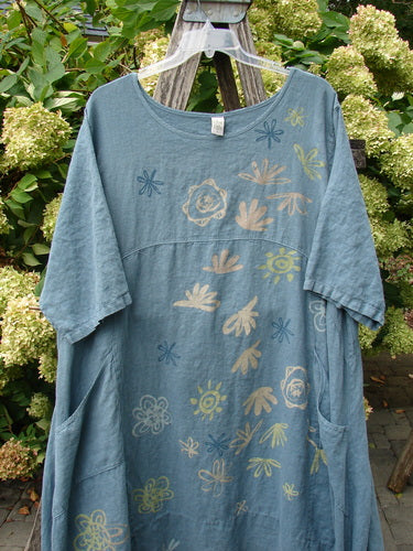 Barclay Linen Lucky Urchin Dress Floral Drift Delaware size 2 in faded blue linen, with a loose short-sleeve silhouette, rounded neckline, and scattered floral drift motifs in pale yellow, cream, and navy. It hangs on a clear hanger against dense green foliage.