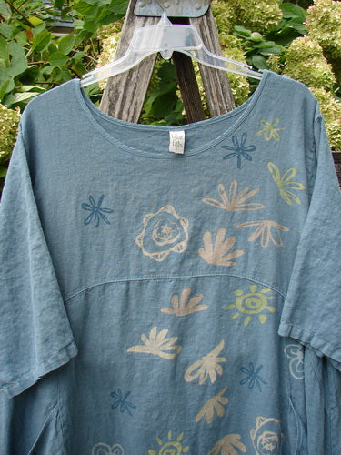 Barclay Linen Lucky Urchin Dress Floral Drift Delaware size 2 in faded blue linen, with a wide scoop neckline and short sleeves. Soft floral and shell-like motifs scatter across the front, hanging from a clear plastic hanger against leafy greenery and weathered wood.