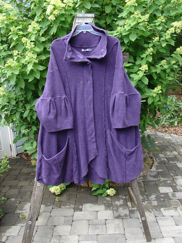 Barclay Patched Celtic Moss Double Snap Button Back Coat in violet hangs on a ladder, showcasing its dropped shoulders, elbow-length pleated sleeves, oversized pockets, and A-line flair, embodying cozy elegance.
