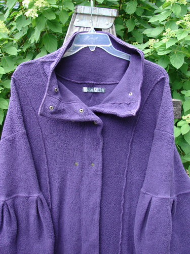 Barclay Patched Celtic Moss Double Snap Button Back Coat in Violet, featuring a high collar, snap closures, oversized pockets, and pleated sleeves, hangs outdoors on a clear plastic hanger.