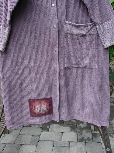 Barclay Patched Chenille Straight Coat Aubergine Dust Size 1, draped over a wooden bench, features a button-front design, large patch pockets, decorative fabric patches, and cozy sleeves with rollable lowers.