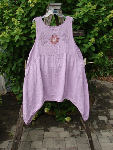 Barclay Linen Tuesday's Pocket Prairie Dress Celtic Floral Lilac Size 1, featuring floral embroidery, pleated empire waist, and diagonal pockets, displayed on a wooden ladder against a leafy patio backdrop.