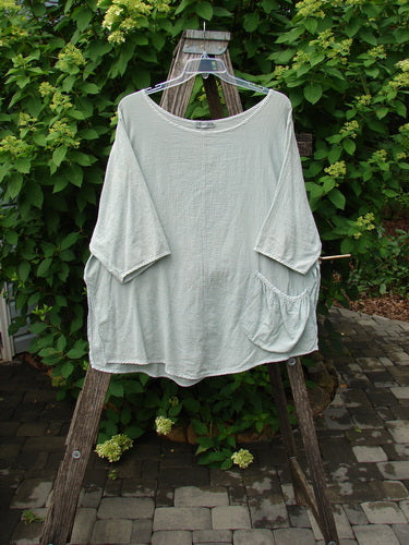 Barclay Gauze Round Bottom Tie Back Pocket Top Unpainted Grass Size 2, displayed on a clear hanger over a wooden ladder, showcasing its loose fit, elbow-length sleeves, and rounded neckline with a single patch pocket.