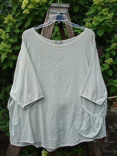 Barclay Gauze Round Bottom Tie Back Pocket Top Unpainted Grass Size 2 displayed on a clear hanger over a wooden ladder, highlighting its loose fit and three-quarter sleeves against a backdrop of green foliage.