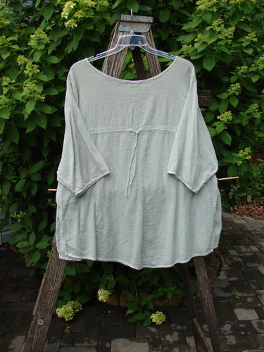 Barclay Gauze Round Bottom Tie Back Pocket Top Unpainted Grass Size 2 draped on a translucent hanger over a wooden stepladder, set against lush hydrangea shrubs, showcasing its relaxed, artistic style.
