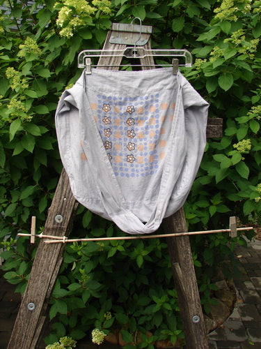 Barclay Linen Sling Tote Daisy Row Lavender OSFA displayed on a ladder, showcasing its wide shoulder sling and slouchy look with painted daisies, against a lush green hedge background.