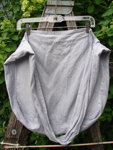 Barclay Linen Sling Tote Daisy Row Lavender OSFA, a trapezoidal light-gray linen piece on a hanger, showcased outdoors. Features include wide shoulder sling, slouchy look, rounded bottom, and daisy row theme.