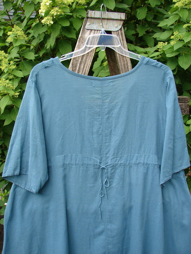Barclay Batiste Flutter Bib Tie Back Dress Honeycomb Tealen Size 1 shown hanging on a wooden ladder, highlighting its loose fit and drawstring tie at the waist, set against lush greenery.