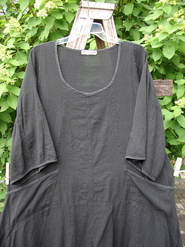 Barclay Gauze Urchin Bell Dress Unpainted Black Size 1: A loose-fitting, long tunic-style dress with elbow-length sleeves and patch pockets, displayed outdoors on a wooden ladder amidst lush greenery.
