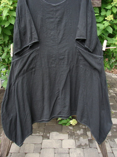 Barclay Gauze Urchin Bell Dress Unpainted Black Size 1, featuring a textured, loose fit, scooped neckline, elbow-length sleeves, large side pockets, and an asymmetrical, drapey hem, displayed on an outdoor wooden rack.