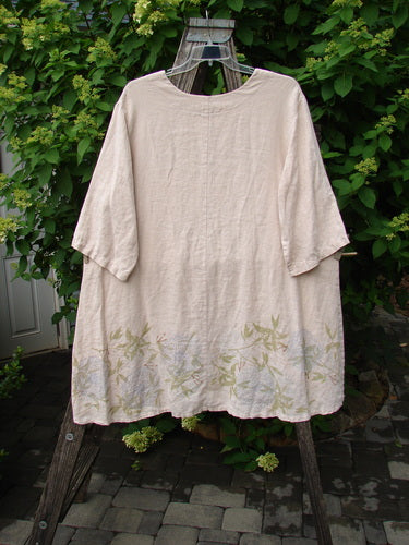 Barclay Linen Hi Low Tunic Top Bamboo Border Plantain Size 2, a loose-fitting natural-linen blouse with elbow-length sleeves, features a hand-painted leaf motif along the hem, hanging outdoors on a wooden stand.