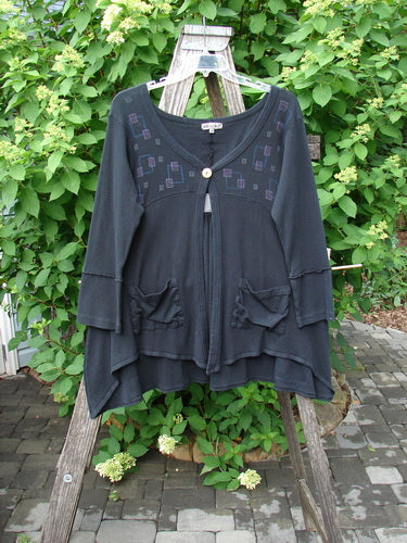 Barclay Patched Thermal Single Button Cardigan Tiny Square Black Size 0 hangs on a ladder, showcasing its scooped neckline, geometric embroidery, and asymmetrical hem against a lush green backdrop.