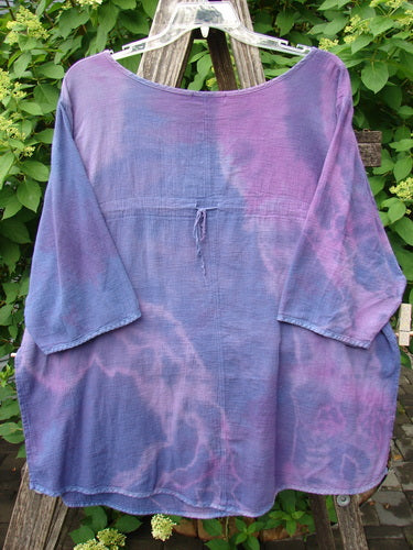 Barclay Gauze Round Bottom Tie Back Pocket Top Mottled Purple Dusk Size 2, a loose-fitting, long-sleeved blouse with tie-dye patterns, displayed on a hanger over a wooden ladder.