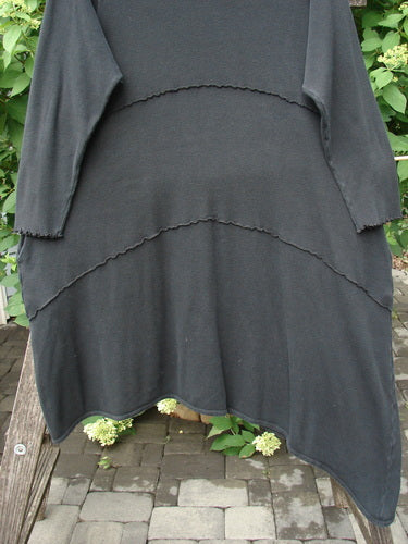 Barclay Thermal Exterior Stitch Three Tier Dress Unpainted Black Size 2 displayed outdoors, showcasing its long sleeves and asymmetrical, lightly ruffled hem tiers against a backdrop of paving stones and hydrangeas.