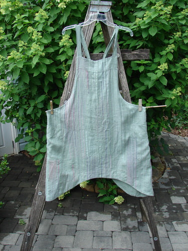 Barclay Linen Side Pocket Apron Jumper Verticals Sage Size 0 displayed in a garden, showcasing its bib front, side ties, adjustable straps, banded upper bib, and wrap drop side pockets.