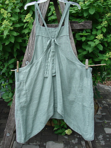 Barclay Linen Side Pocket Apron Jumper Verticals Sage Size 0 features crisscross tie-back straps and wide front pockets, crafted from lightweight linen, hanging on an outdoor wooden frame.
