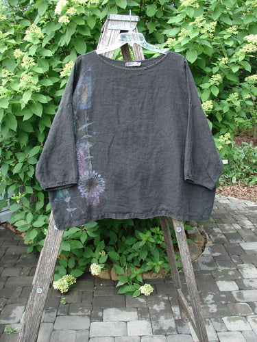 Barclay Linen Lantern Boxy Top Side Floral Black Size 1: A loose-fitting, dark charcoal tunic with a hand-painted floral motif on one side, displayed draped over a wooden ladder, showcasing its unique, artistic design.